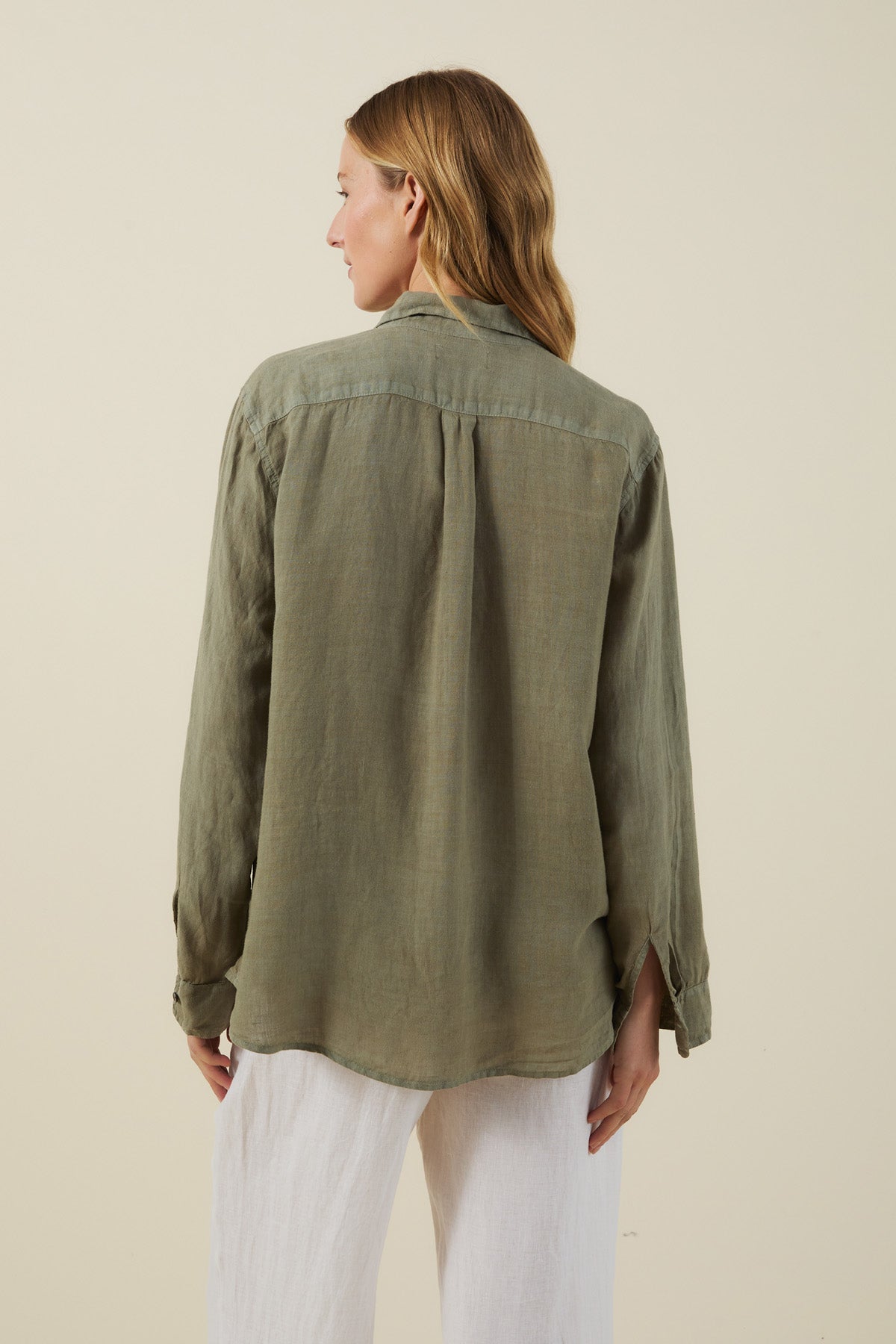 A woman with light brown hair faces away from the camera, wearing the MULHOLLAND LINEN SHIRT by Velvet by Jenny Graham in olive green, paired with white pants against a neutral background.-41645087260865