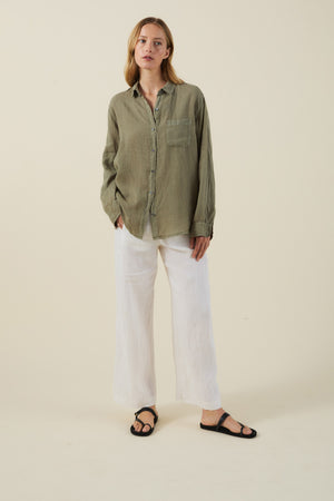 A person stands against a plain background wearing the MULHOLLAND LINEN SHIRT by Velvet by Jenny Graham in olive green, styled with white wide-leg pants and black sandals.