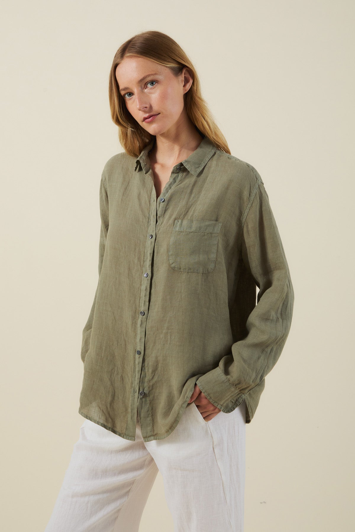 A woman wears the MULHOLLAND LINEN SHIRT by Velvet by Jenny Graham—an olive green, breathable linen long-sleeve with a chest pocket—paired with white pants, against a plain light background.-41645087457473