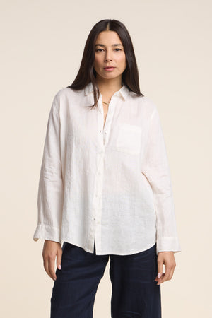 A woman models the MULHOLLAND LINEN SHIRT by Velvet by Jenny Graham—a loose, long-sleeve linen top with a chest pocket—paired with dark pants, standing against a plain light background.