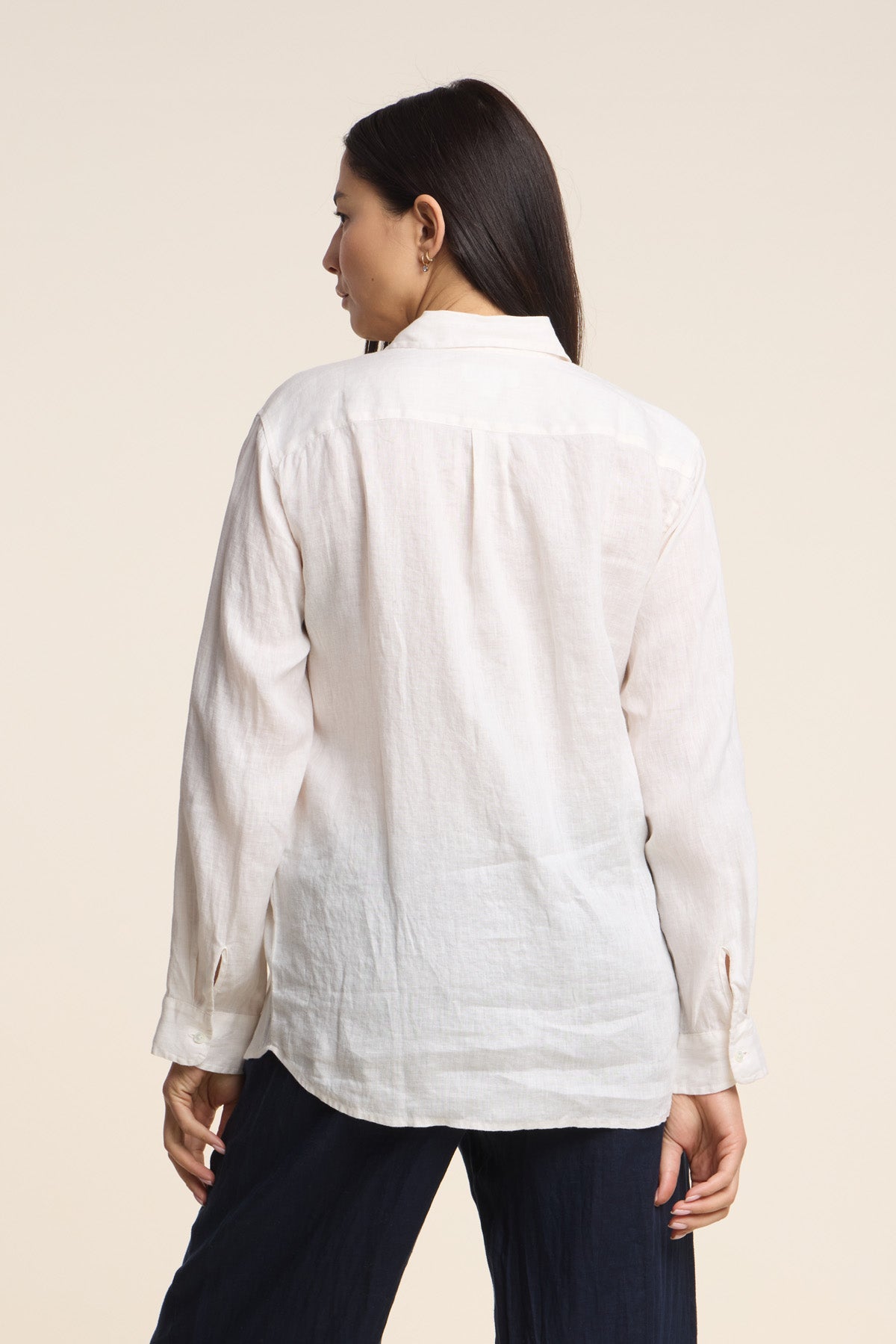 A woman, seen from behind, wears the MULHOLLAND LINEN SHIRT by Velvet by Jenny Graham—a breathable, light-colored button-up—paired with dark pants against a plain background.-41476546592961
