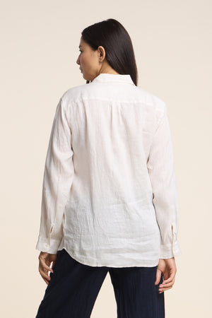 A woman, seen from behind, wears the MULHOLLAND LINEN SHIRT by Velvet by Jenny Graham—a breathable, light-colored button-up—paired with dark pants against a plain background.
