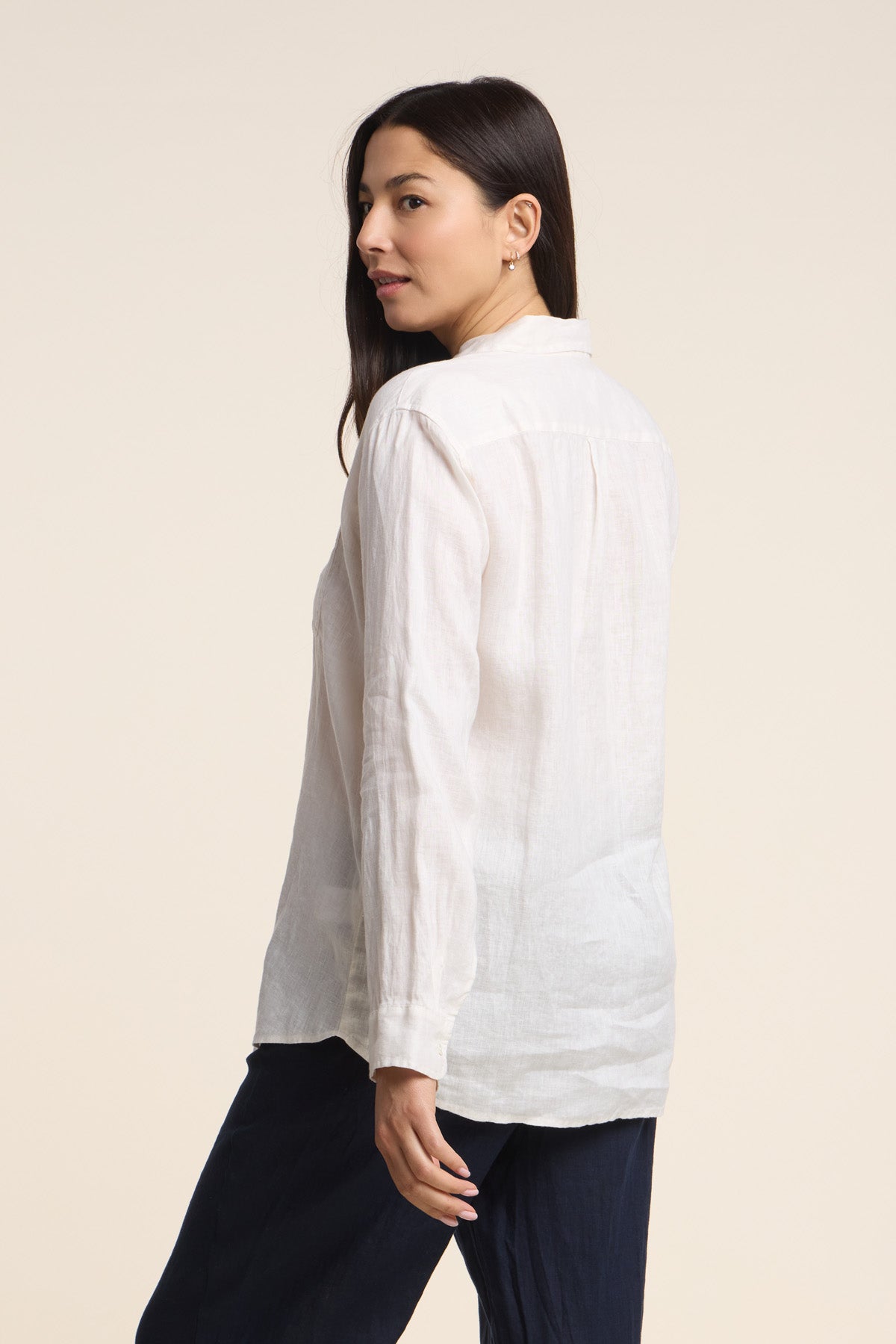 A woman faces away, turning left, wearing the MULHOLLAND LINEN SHIRT by Velvet by Jenny Graham—a long-sleeve white button-up—paired with dark pants against a neutral background.-41476546560193