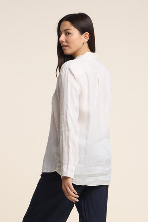 A woman faces away, turning left, wearing the MULHOLLAND LINEN SHIRT by Velvet by Jenny Graham—a long-sleeve white button-up—paired with dark pants against a neutral background.