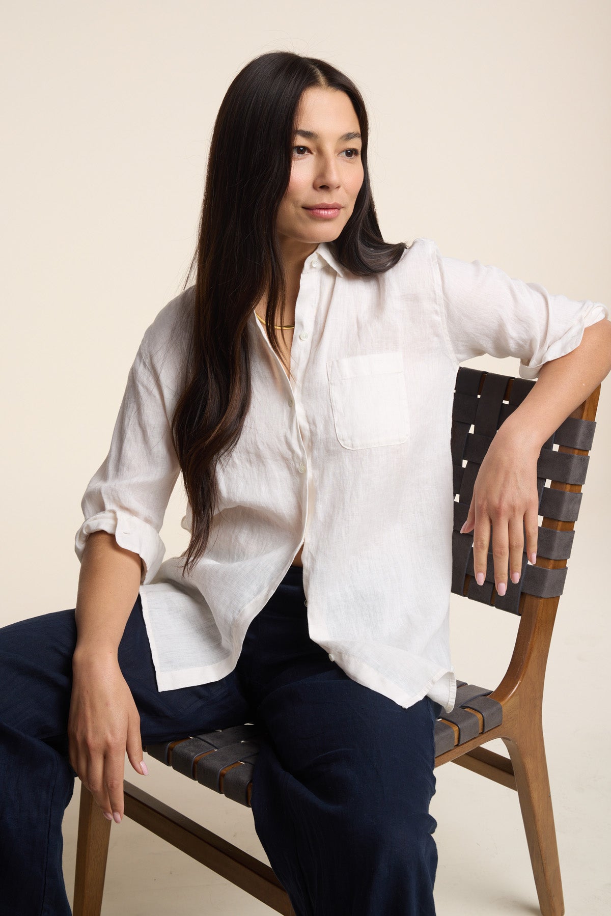 A woman with long dark hair wears the MULHOLLAND LINEN SHIRT by Velvet by Jenny Graham and dark pants, seated on a wooden chair and gazing slightly to the side with a neutral expression.