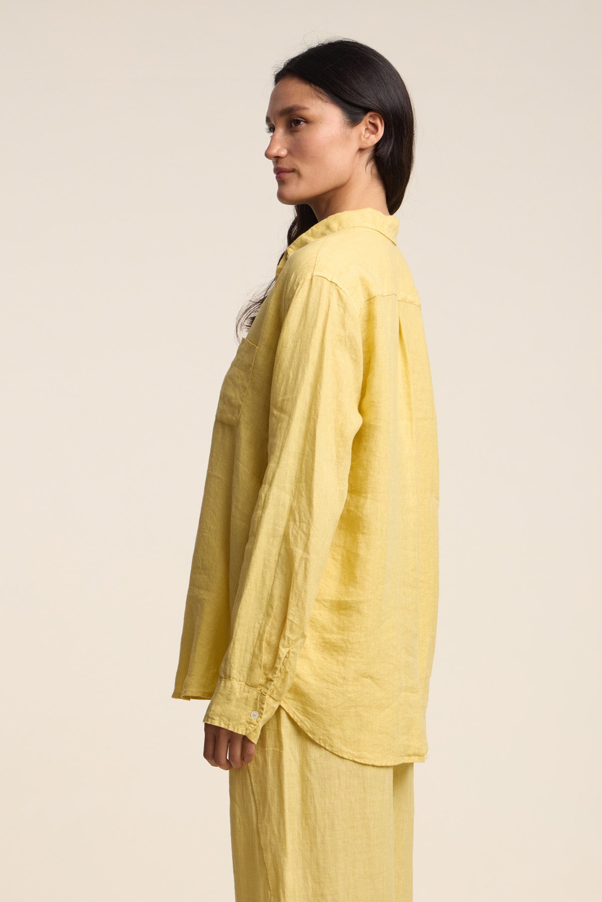 A woman stands in profile against a plain light background, wearing the MULHOLLAND LINEN SHIRT by Velvet by Jenny Graham, a loose-fitting, long-sleeved yellow woven linen top styled with matching yellow pants.