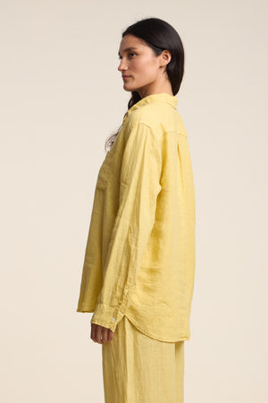 A woman stands in profile against a plain light background, wearing the MULHOLLAND LINEN SHIRT by Velvet by Jenny Graham, a loose-fitting, long-sleeved yellow woven linen top styled with matching yellow pants.