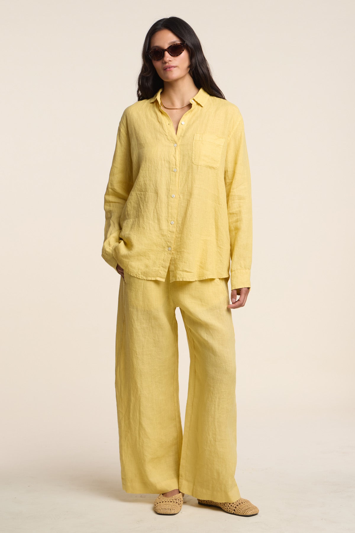 A woman wears Velvet by Graham & Spencer’s LOLA LINEN PANT with a loose yellow linen button-up shirt, sunglasses, and woven slip-on shoes.