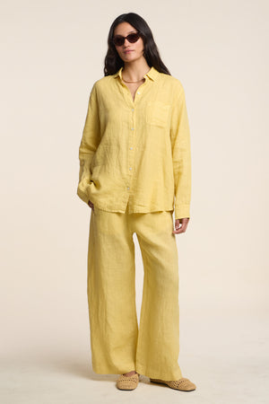A woman wears Velvet by Graham & Spencer’s LOLA LINEN PANT with a loose yellow linen button-up shirt, sunglasses, and woven slip-on shoes.
