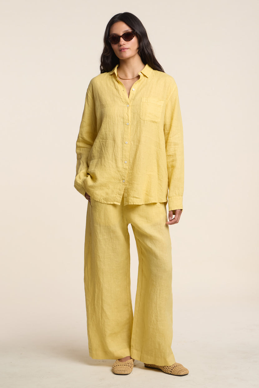 A woman wears Velvet by Graham & Spencer’s LOLA LINEN PANT with a loose yellow linen button-up shirt, sunglasses, and woven slip-on shoes.