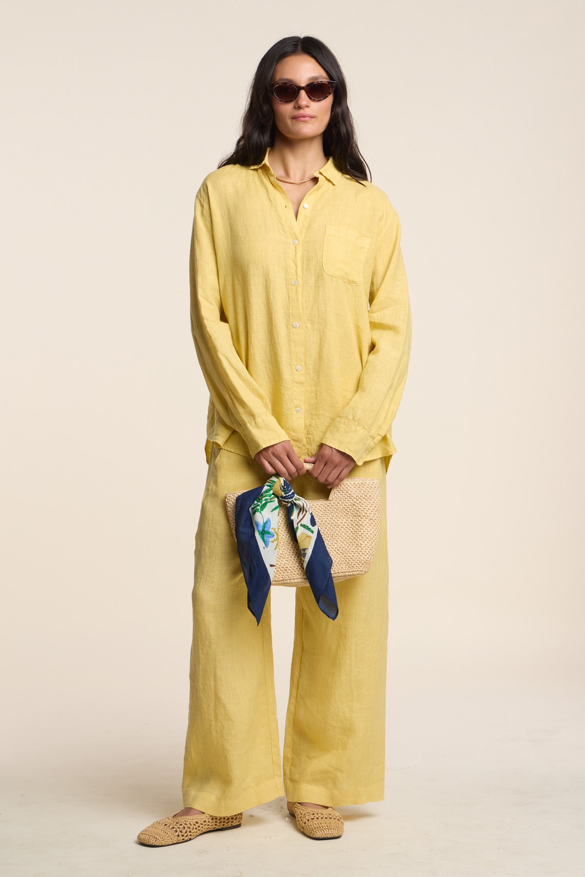 Woman in a yellow linen shirt and the LOLA LINEN PANT by Velvet by Graham & Spencer—relaxed, straight-leg, cropped pants—wearing sunglasses, holding a woven clutch with a floral scarf, and standing against a plain beige background.