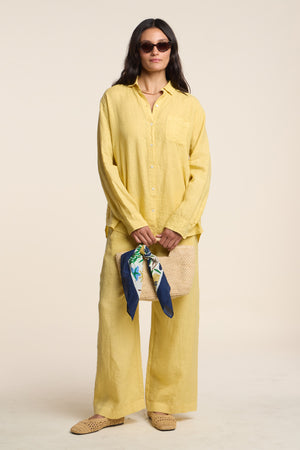Woman in a yellow linen shirt and the LOLA LINEN PANT by Velvet by Graham & Spencer—relaxed, straight-leg, cropped pants—wearing sunglasses, holding a woven clutch with a floral scarf, and standing against a plain beige background.