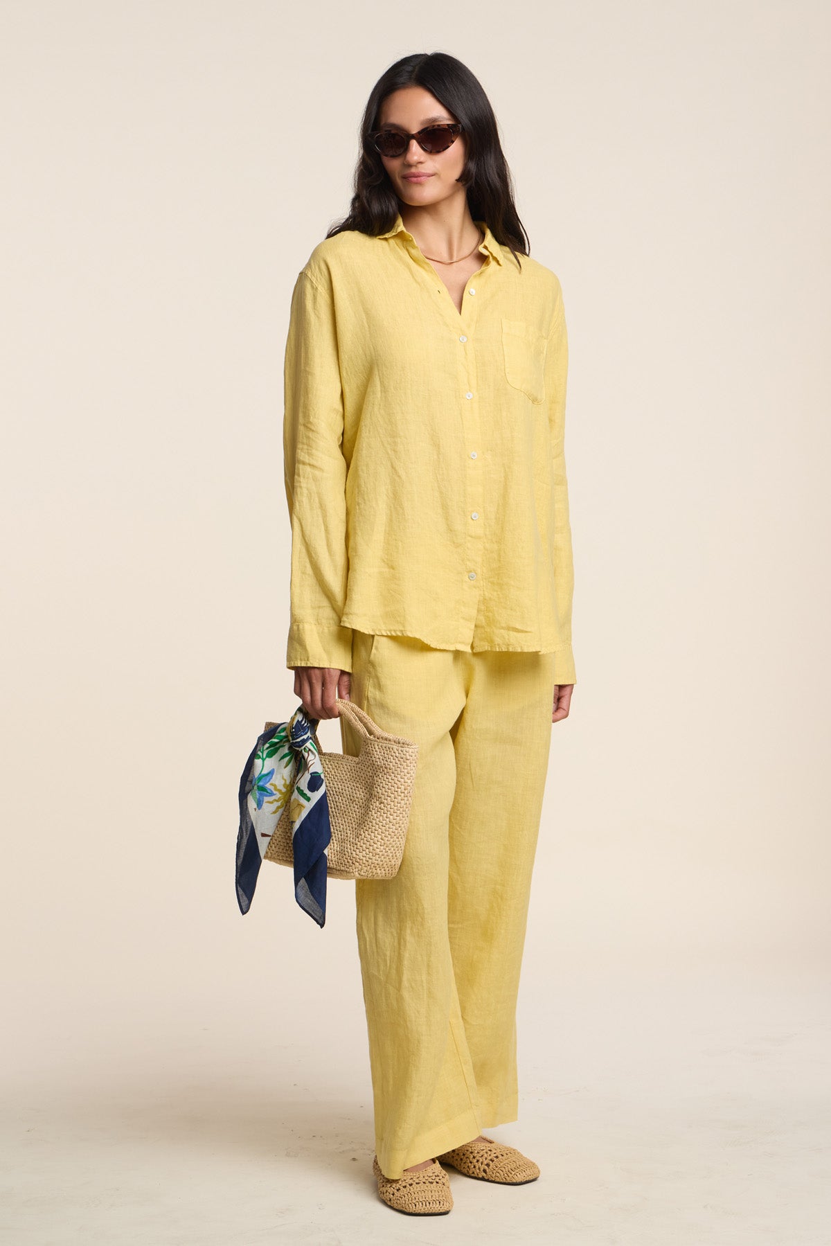 A woman wears the Velvet by Graham & Spencer LOLA LINEN PANT in yellow with a matching loose button-up shirt, accessorized with sunglasses, flat shoes, and a woven bag topped with a scarf.-41527072784577