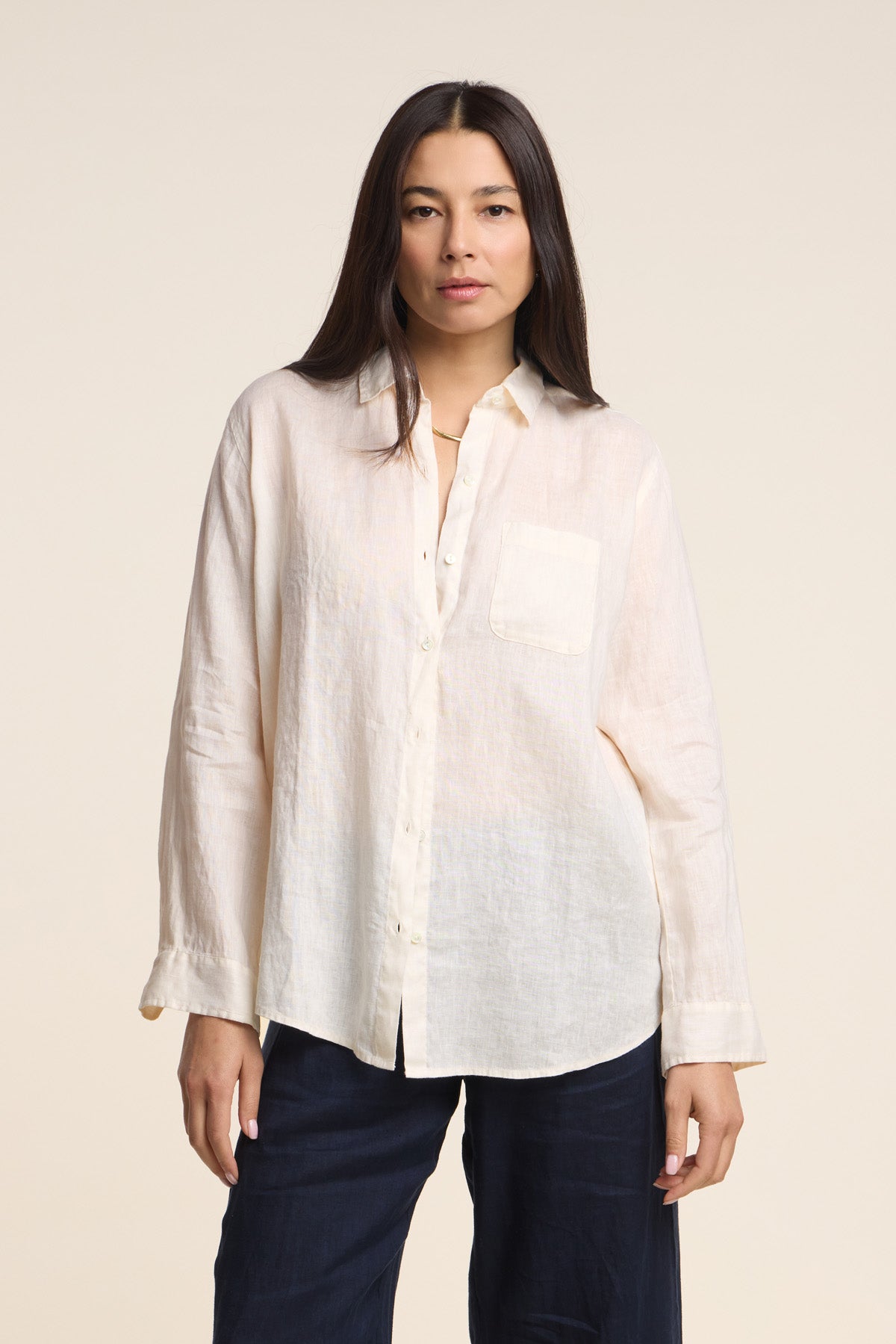 A woman stands against a plain background wearing the MULHOLLAND LINEN SHIRT by Velvet by Jenny Graham, a loose, cream-colored woven linen shirt with a front pocket, paired with dark pants.-41637040750785
