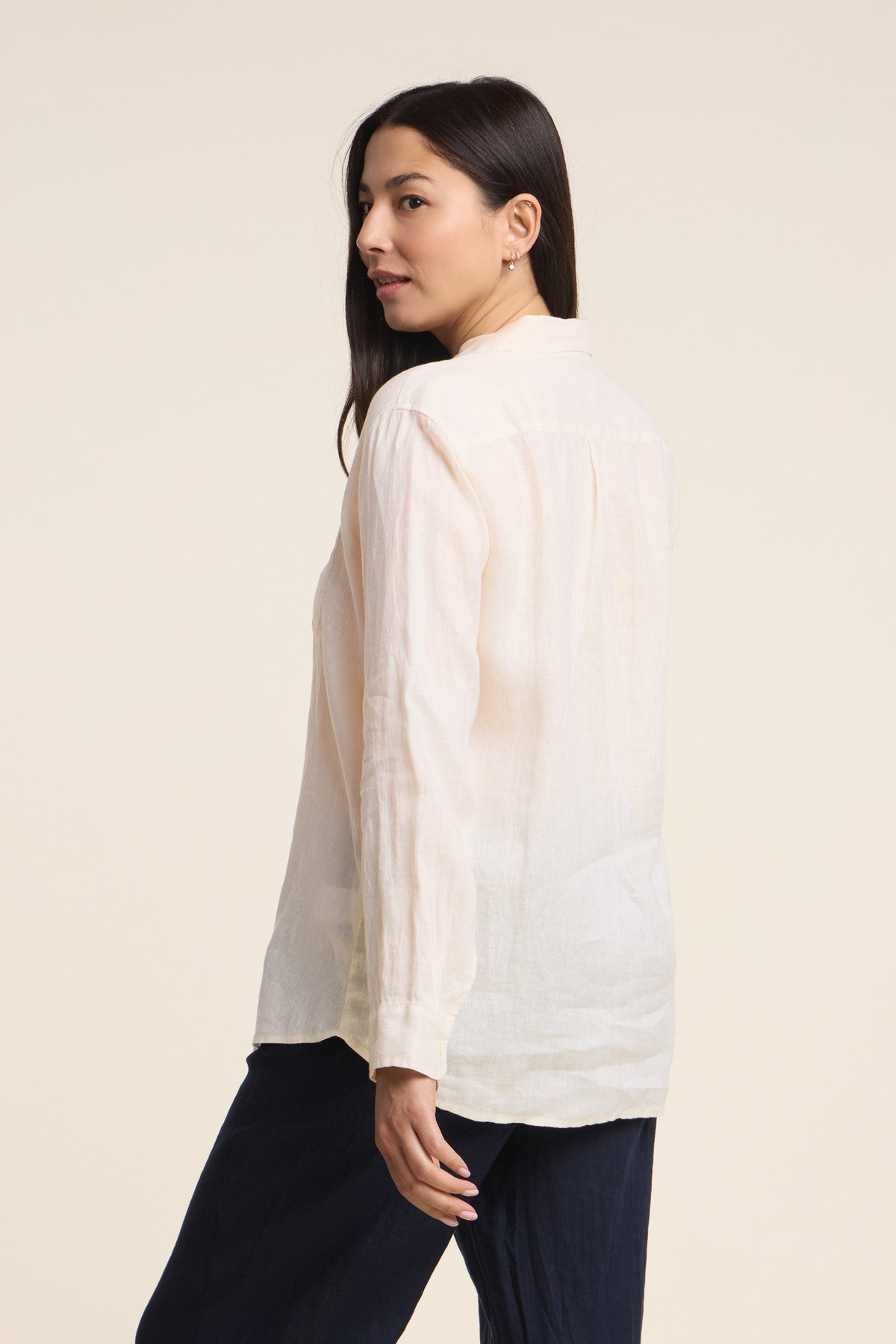 A woman stands sideways facing away, wearing the MULHOLLAND LINEN SHIRT by Velvet by Jenny Graham—a lightweight linen button-up with a relaxed fit—paired with dark pants, set against a plain beige background.-41637040685249