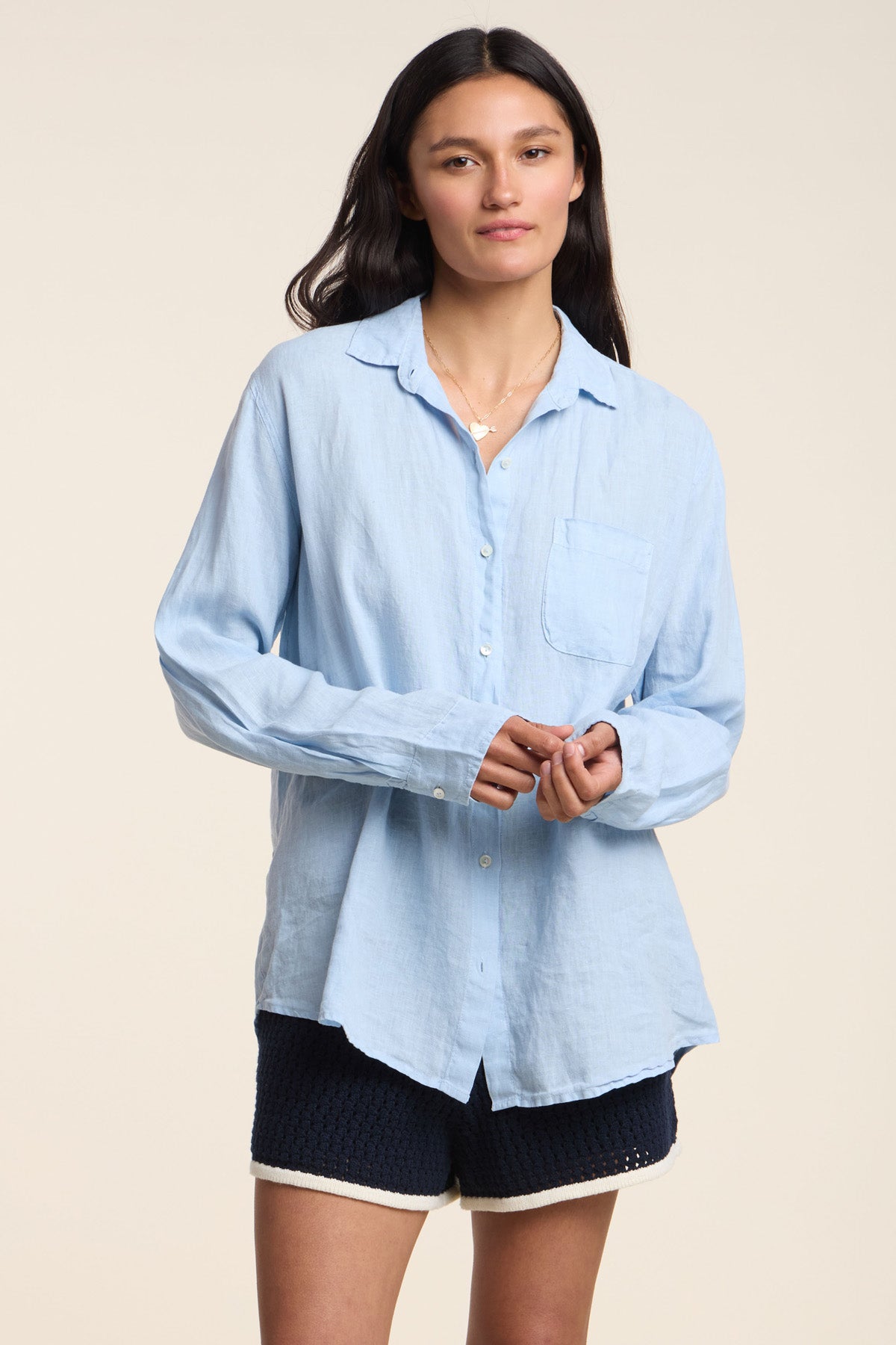 A person stands facing forward, wearing the MULHOLLAND LINEN SHIRT by Velvet by Jenny Graham—a light blue button-up with a chest pocket—and dark shorts, set against a plain light background.