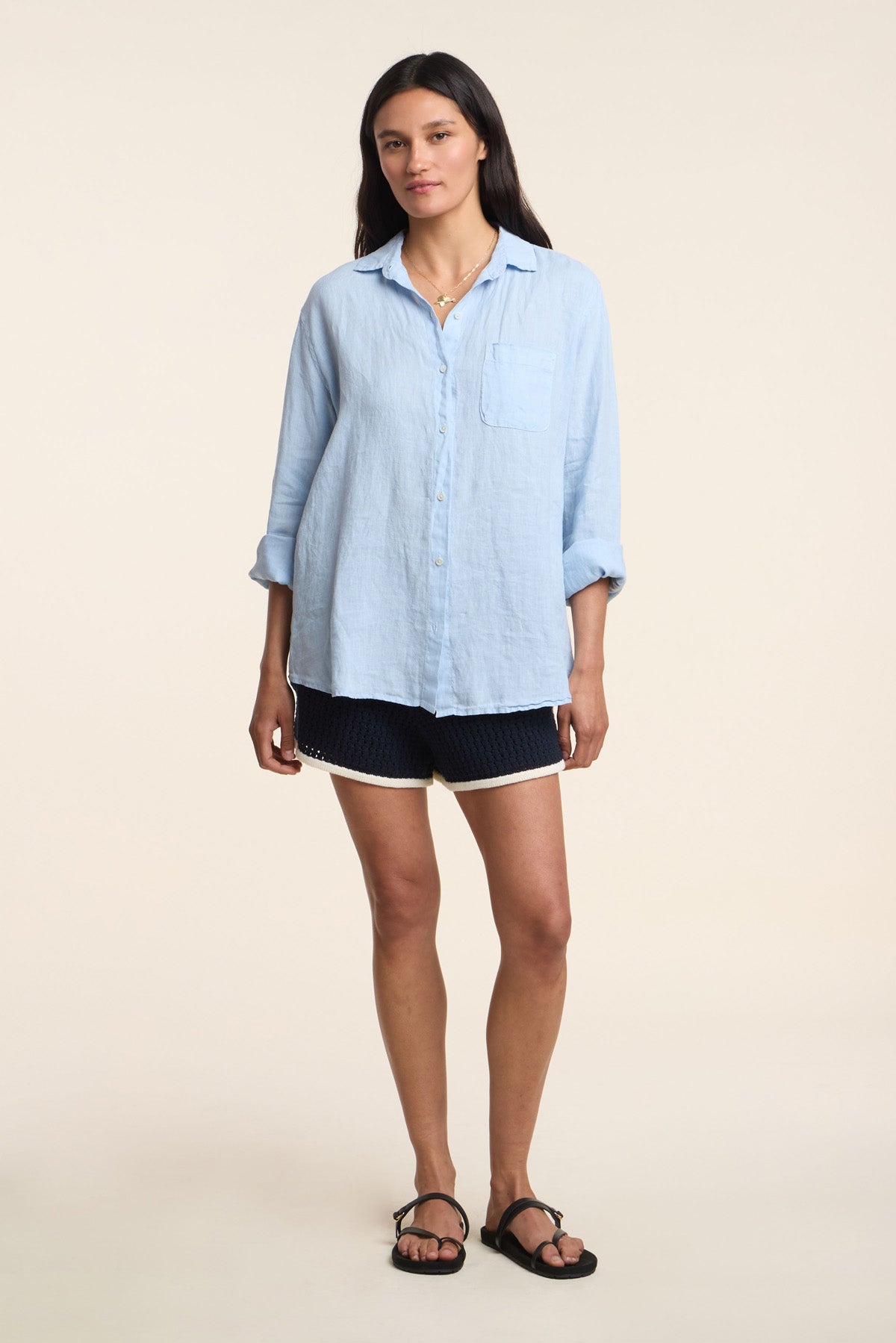 A woman stands facing forward against a plain light background, wearing the MULHOLLAND LINEN SHIRT by Velvet by Jenny Graham—a light blue button-up with a relaxed silhouette—paired with navy shorts featuring white trim and black sandals.