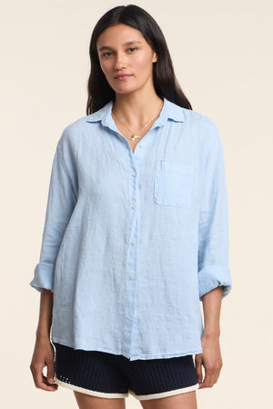 A woman wears the MULHOLLAND LINEN SHIRT by Velvet by Jenny Graham—a light blue, breathable linen button-up with a chest pocket and rolled-up sleeves—paired with black shorts against a plain background.