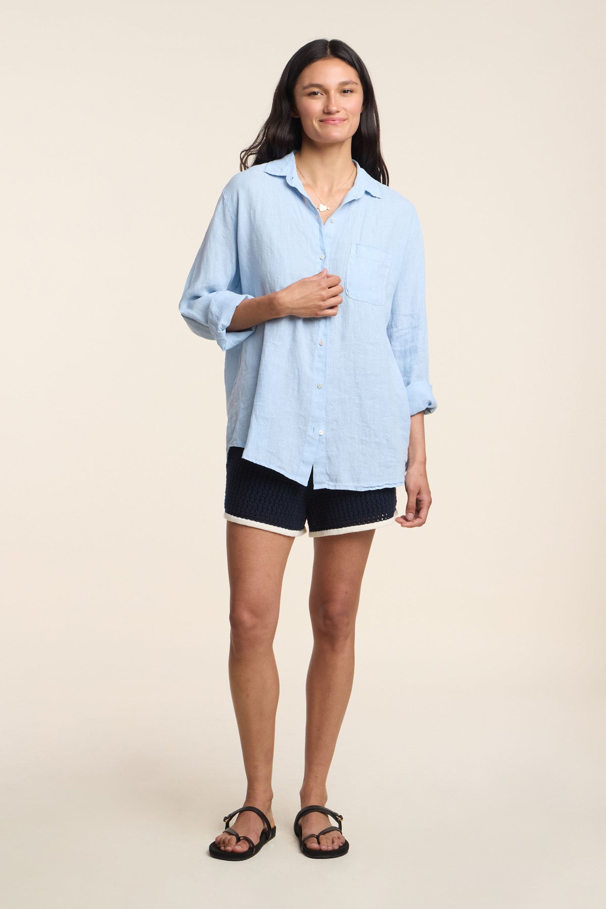A person stands against a plain background wearing a relaxed silhouette: the MULHOLLAND LINEN SHIRT by Velvet by Jenny Graham in light blue, paired with dark shorts with light trim and black sandals.