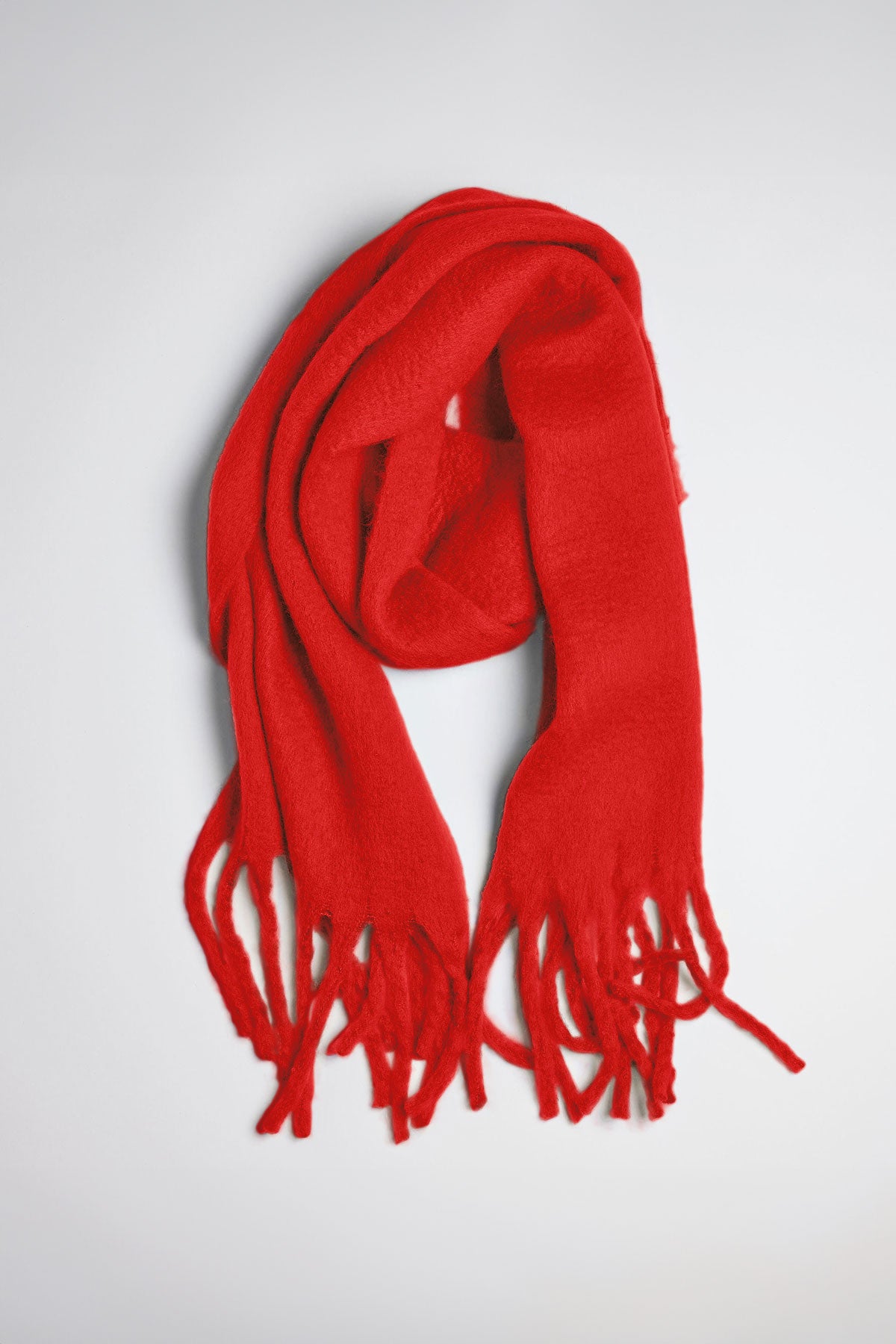   The CHIC SOLID SCARF by Velvet by Graham & Spencer, a vibrant red accessory with fringed ends, is laid flat on a light gray surface. 