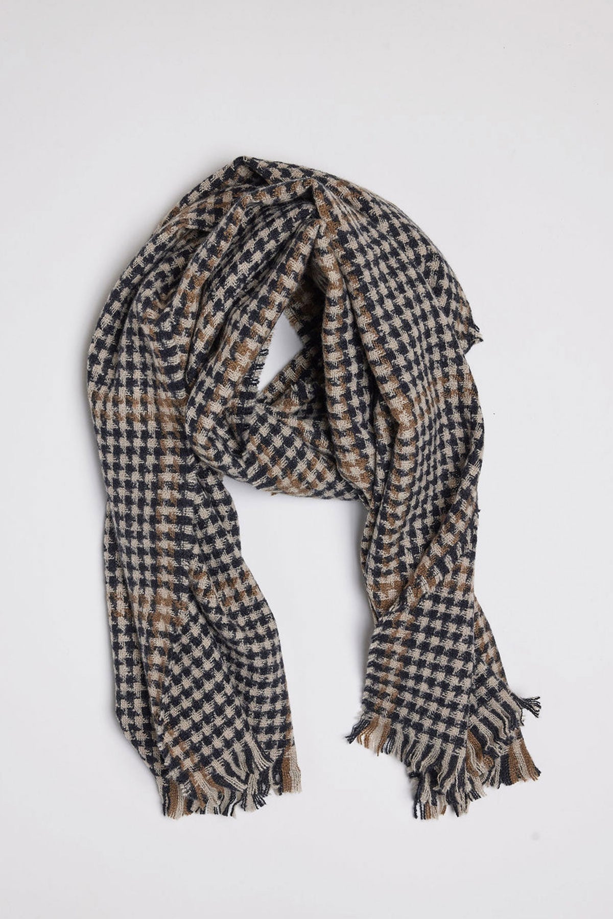   The PEAK SCARF by Velvet by Graham & Spencer, showcasing a houndstooth pattern with fringed edges in a blend of navy blue and beige colors, is displayed flat against a plain light gray background. 