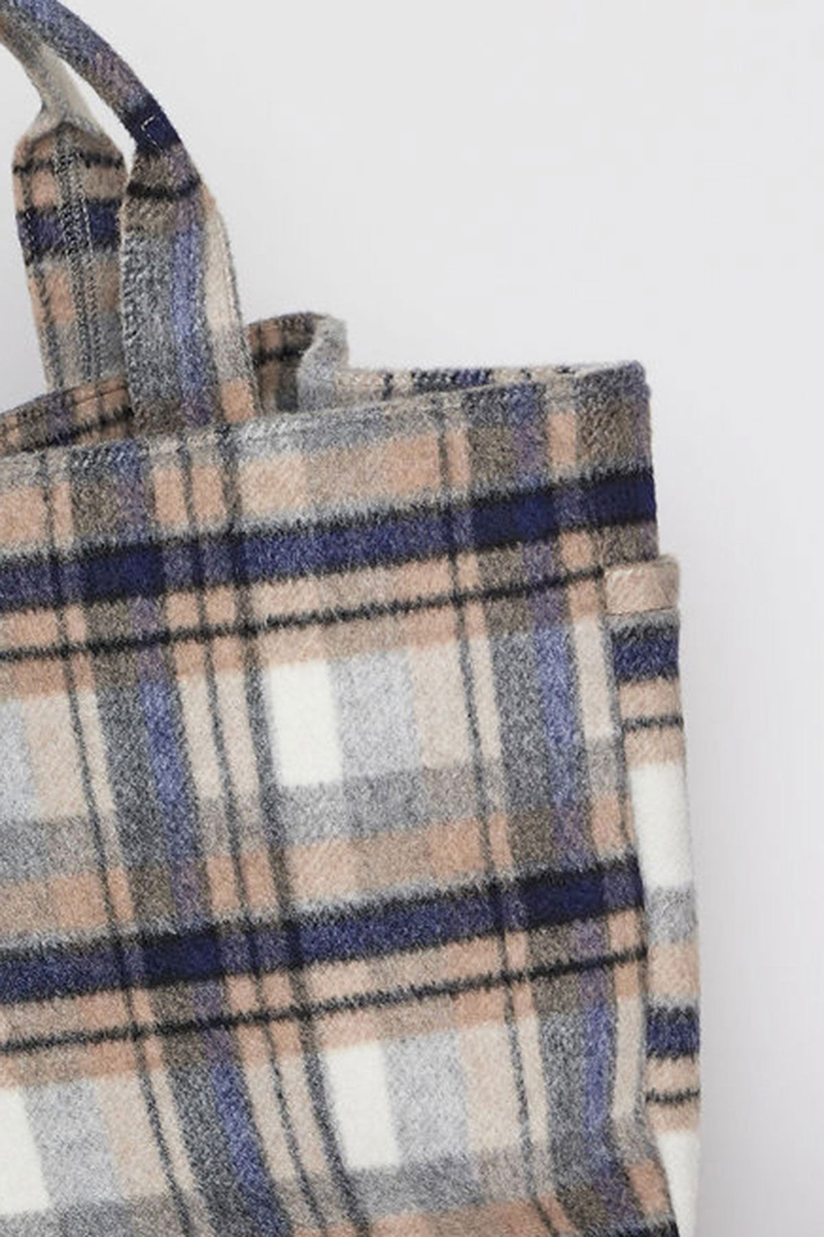   A close-up image of the PLAID TOTE by Velvet by Graham & Spencer showcases its plaid-patterned fabric in shades of blue, gray, white, and beige. The tote bag features dual handles and has a soft texture. 