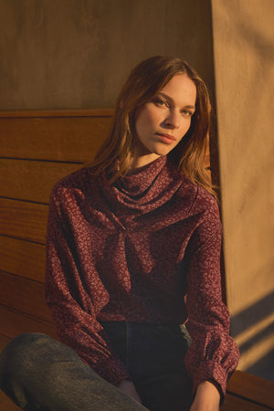 A woman with long brown hair sits on a wooden bench in warm natural light, wearing the Velvet by Graham & Spencer NALA SILK BLOUSE—a cheetah-print silk top featuring a graceful cowl neckline—paired with dark jeans.