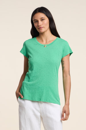 A woman with long dark hair wears the ODELIA TEE by Velvet by Graham & Spencer, a green cotton slub shirt with a classic crew neckline, paired with white pants against a plain background.
