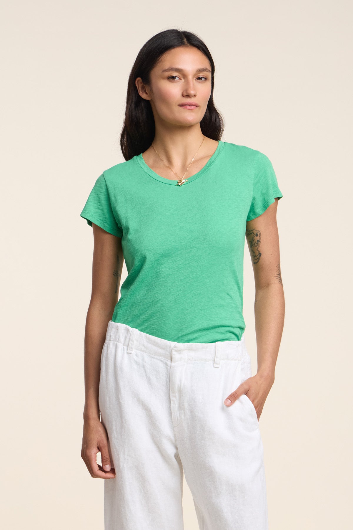A person with long dark hair wears the ODELIA TEE by Velvet by Graham & Spencer—featuring a classic crew neckline in green—with white pants, standing with one hand in their pocket against a plain beige background.