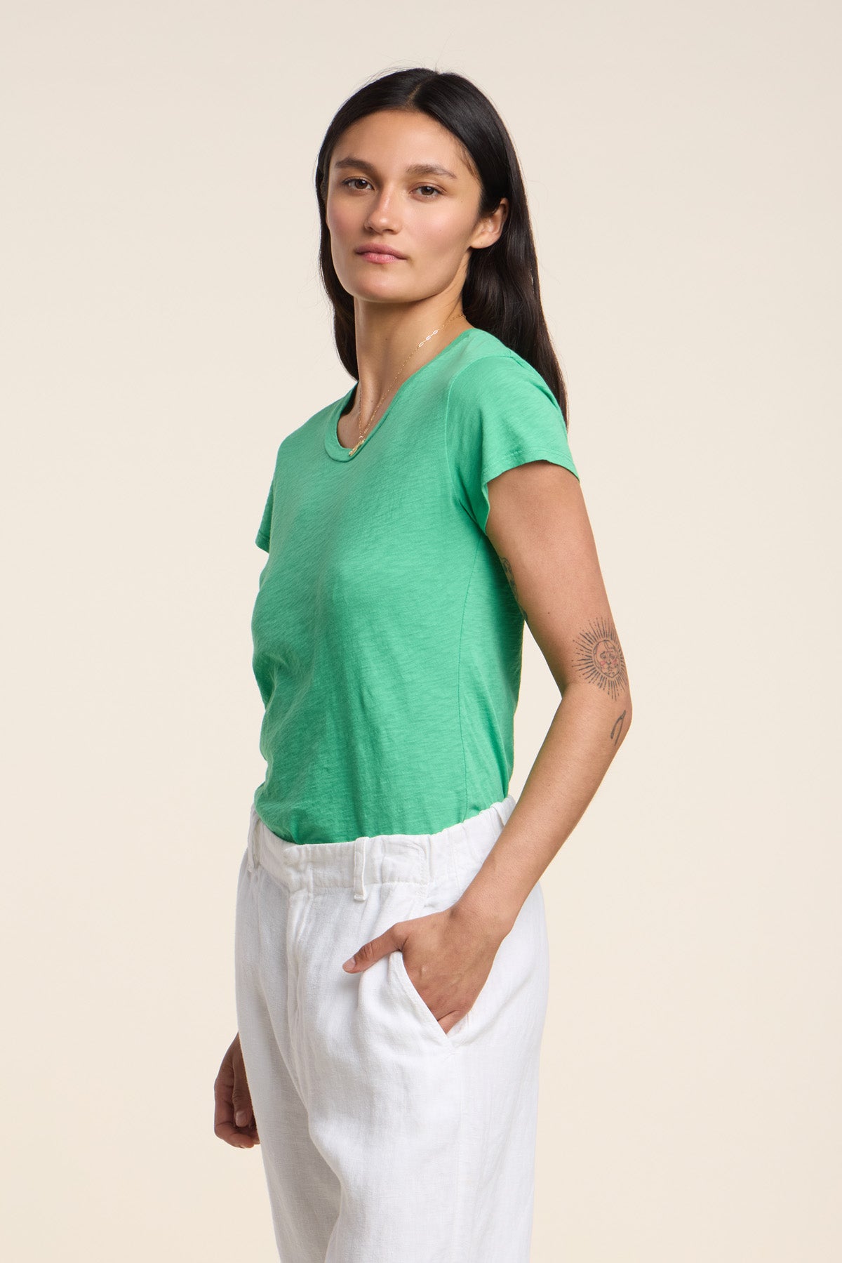 A person with long dark hair wears the ODELIA TEE by Velvet by Graham & Spencer—a green cotton slub crewneck—paired with white pants, standing slightly to the side against a plain background. A true wardrobe essential.