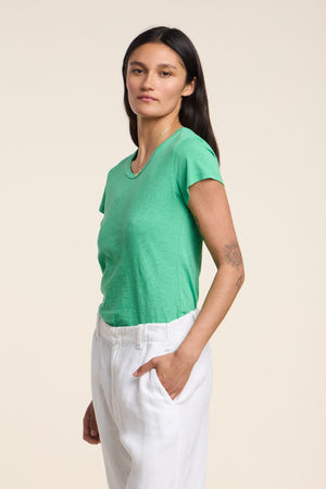 A person with long dark hair wears the ODELIA TEE by Velvet by Graham & Spencer—a green cotton slub crewneck—paired with white pants, standing slightly to the side against a plain background. A true wardrobe essential.