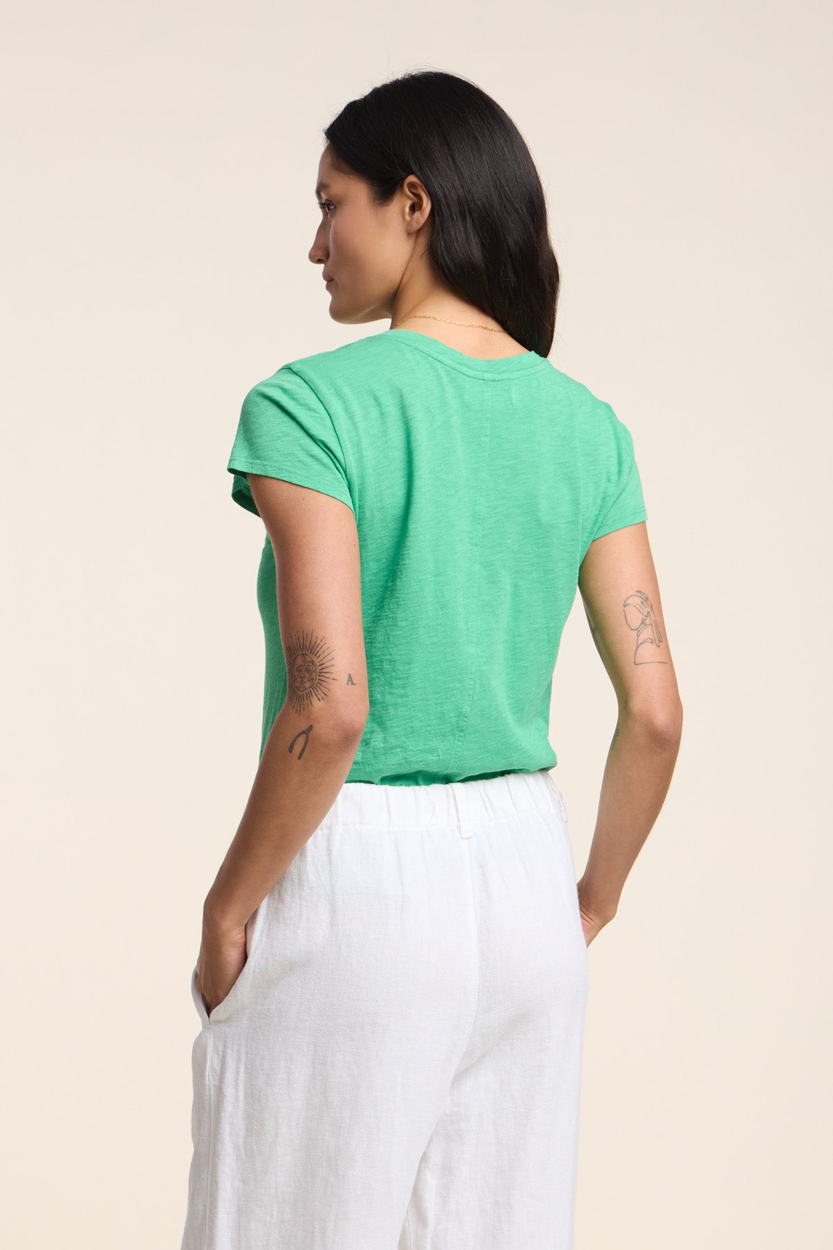 A woman with dark hair, visible tattoos, and white pants stands with her back to the camera, wearing the ODELIA TEE by Velvet by Graham & Spencer—a green cotton slub crew-neck tee that’s a true wardrobe essential.-41527104897217