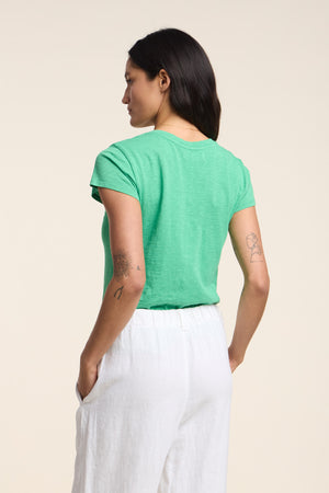 A woman with dark hair, visible tattoos, and white pants stands with her back to the camera, wearing the ODELIA TEE by Velvet by Graham & Spencer—a green cotton slub crew-neck tee that’s a true wardrobe essential.