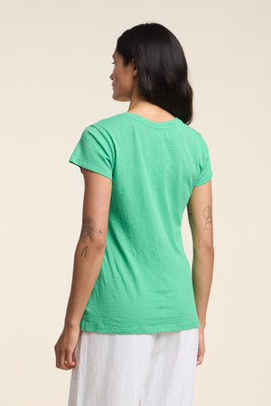 Person wearing the ODELIA TEE by Velvet by Graham & Spencer—a green cotton slub tee with a classic crew neckline—paired with white pants, shown from the back against a plain light background. A wardrobe essential.