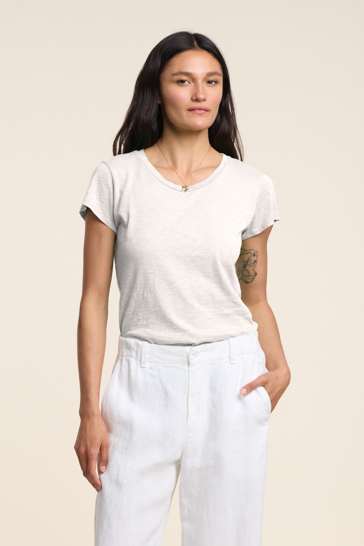 A person with long dark hair wears the ODELIA TEE by Velvet by Graham & Spencer, featuring a classic crew neckline and cotton slub fabric, paired with white pants and posing with one hand in their pocket against a plain background.