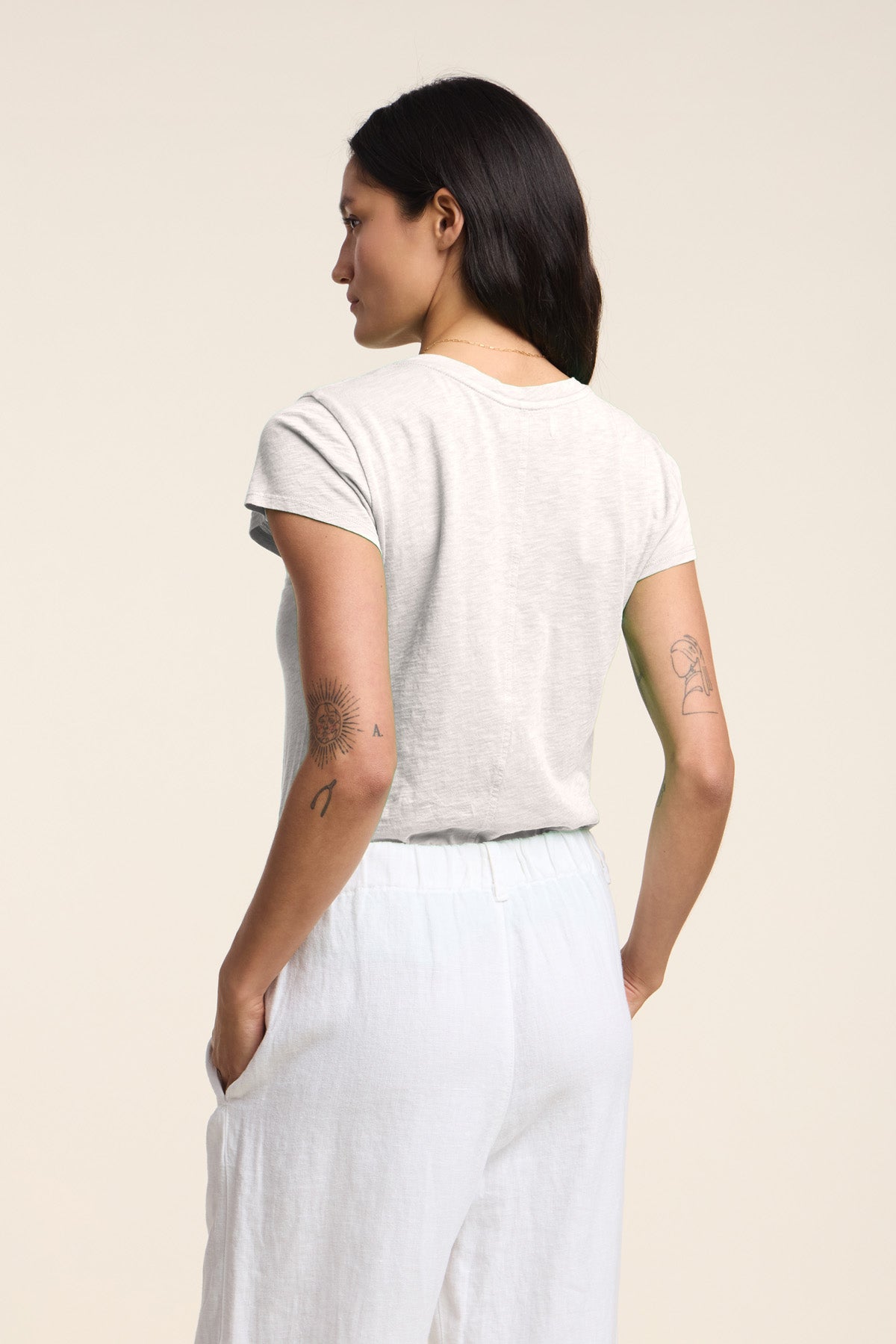 Wearing the Velvet by Graham & Spencer ODELIA TEE, a person with long dark hair and tattoos on both arms stands with their back to the camera—a true wardrobe essential in light gray cotton slub.-41633278165185