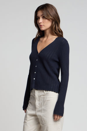 A woman wears the OTTILIE CARDIGAN by Velvet by Graham & Spencer—navy blue, organic cotton-linen, button front—paired with light beige pants against a plain light background.