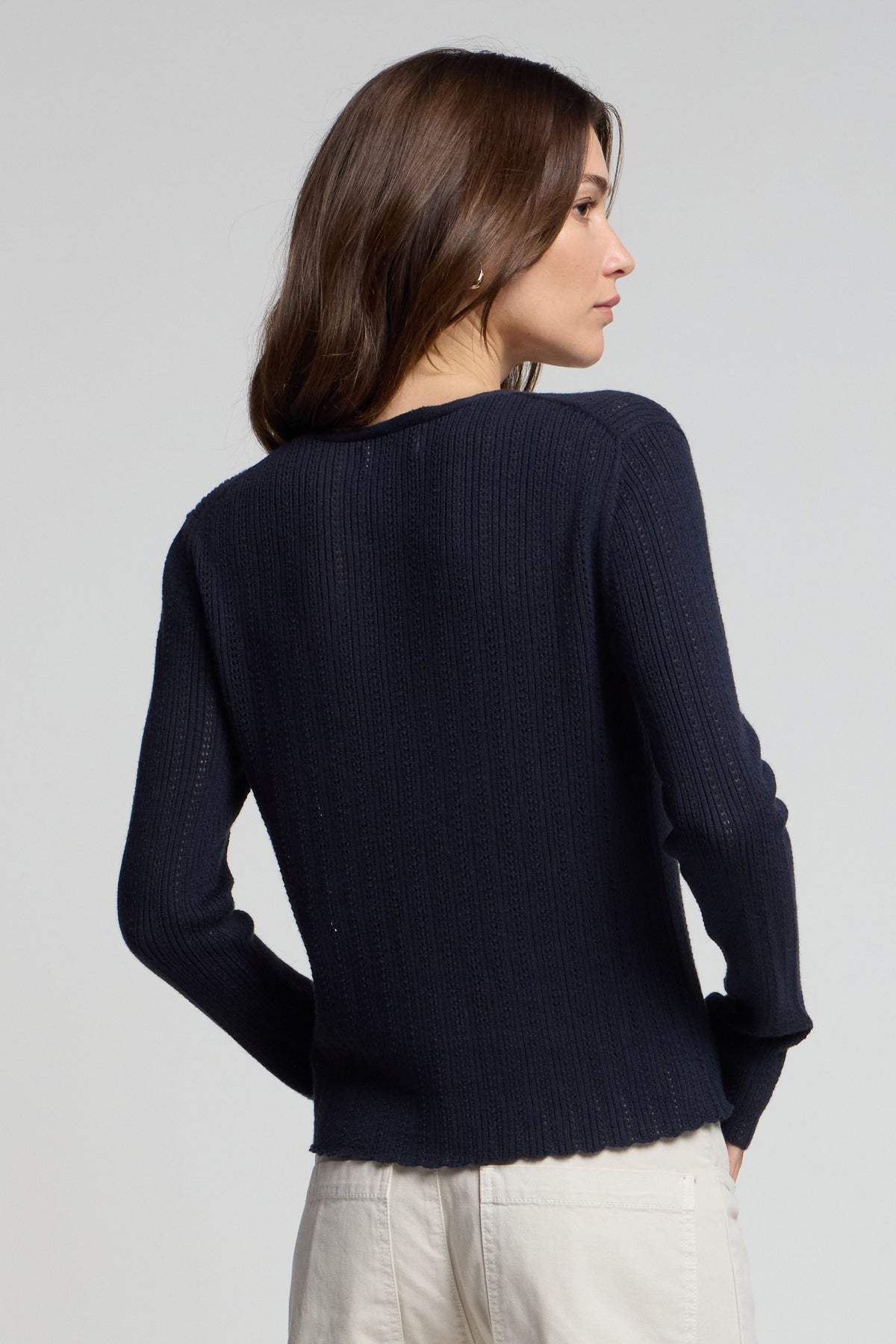 A woman with long brown hair, seen from the back, wears the Velvet by Graham & Spencer OTTILIE CARDIGAN in navy blue organic cotton-linen and light-colored pants.