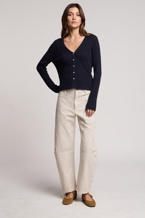 A woman faces forward against a plain light background, wearing the BRYLIE PANT by Velvet by Graham & Spencer, styled with a navy button-up cardigan and brown flats.