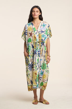A woman faces forward in the PIPPA DRESS by Velvet by Graham & Spencer, a loose, knee-length beach dress with a colorful tropical print and yellow sandals, hands in pockets, against a plain background.