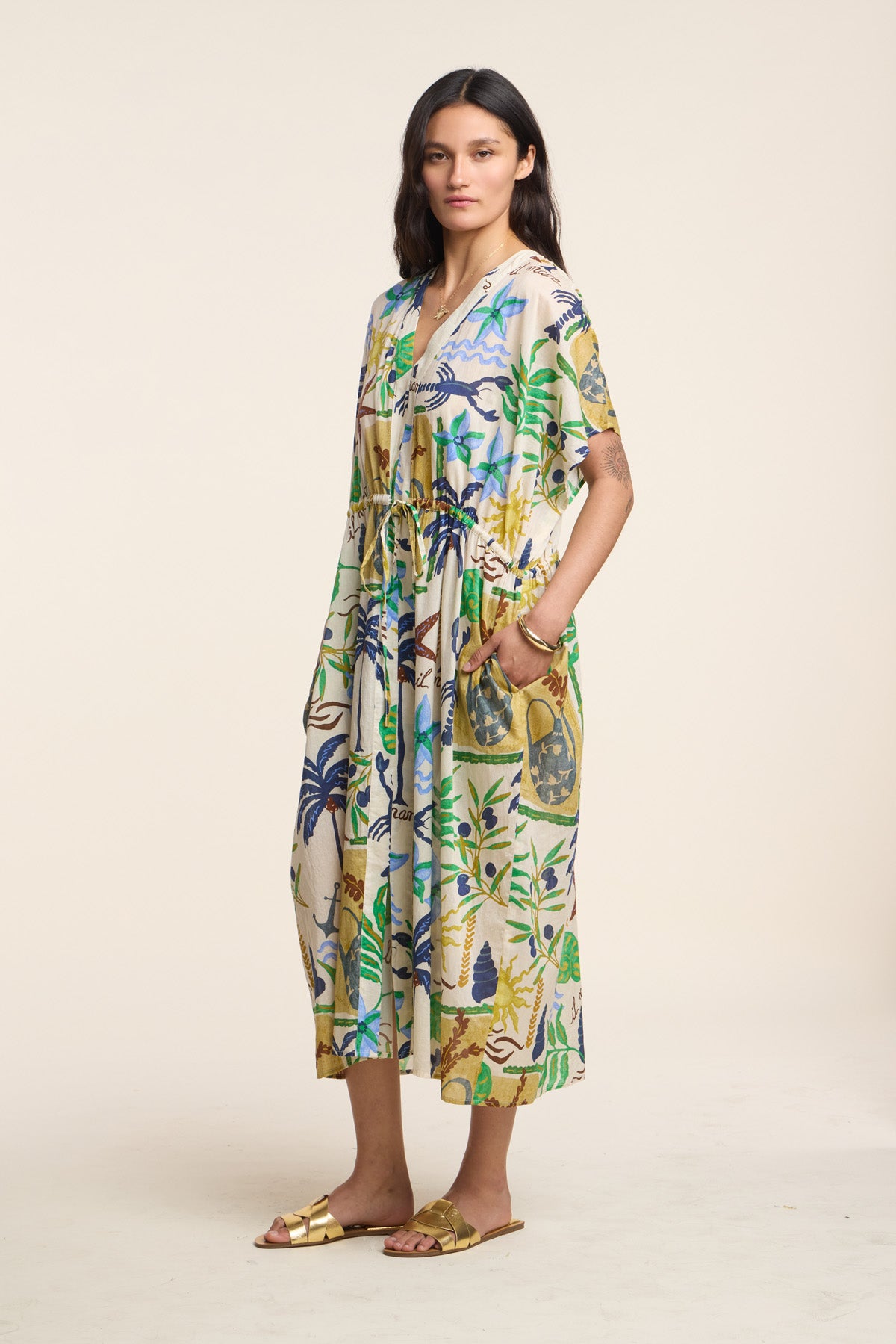 A woman wears the PIPPA DRESS by Velvet by Graham & Spencer, a loose, midi-length dress featuring a colorful botanical print, paired with gold sandals against a plain light background.