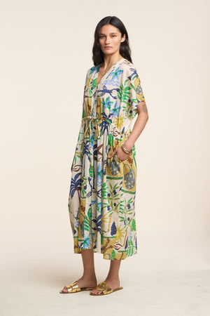 A woman wears the PIPPA DRESS by Velvet by Graham & Spencer, a loose, midi-length dress featuring a colorful botanical print, paired with gold sandals against a plain light background.
