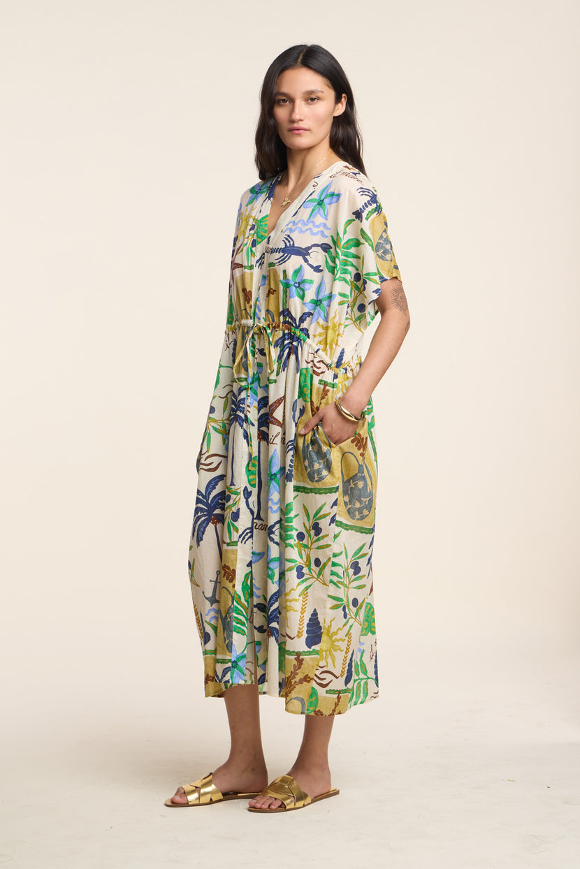 A woman wears the PIPPA DRESS by Velvet by Graham & Spencer, a loose, midi-length dress featuring a colorful botanical print, paired with gold sandals against a plain light background.