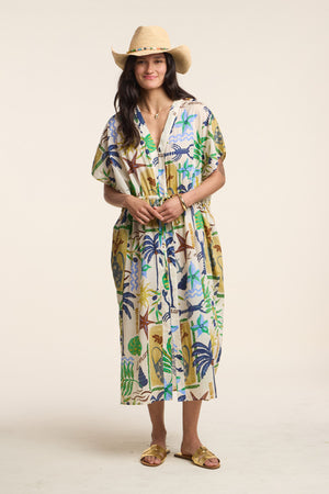 A woman in the Velvet by Graham & Spencer PIPPA DRESS, a straw hat, and yellow sandals faces the camera, her effortlessly chic style highlighted against a plain backdrop.