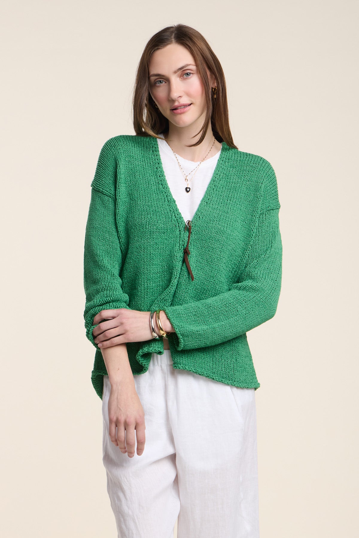 A woman models the Velvet by Graham & Spencer QUINNLY CARDIGAN, a green open-front design in cotton tape yarn, layered over a white top and pants with arms crossed against a neutral background.
