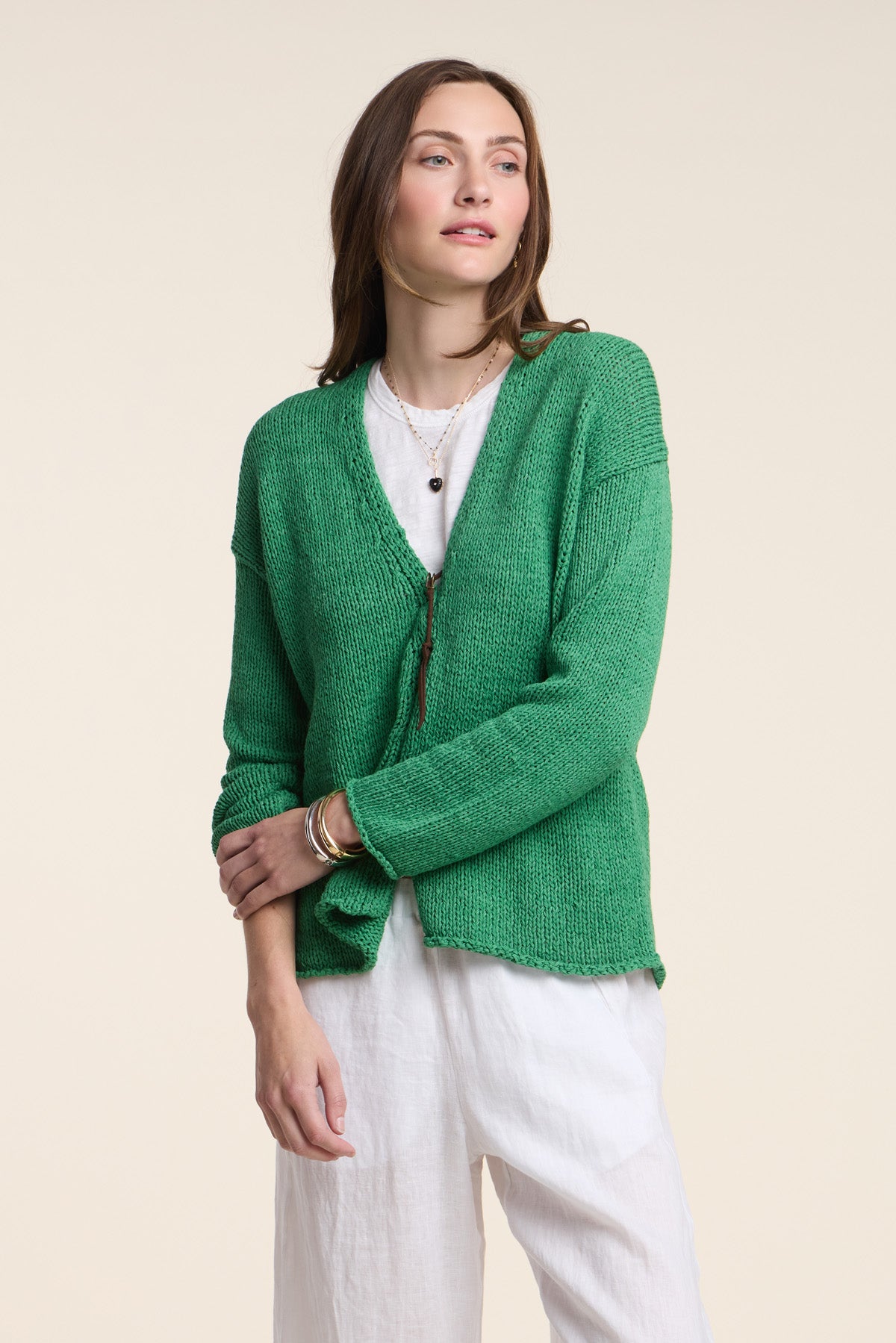 A woman wears the QUINNLY CARDIGAN by Velvet by Graham & Spencer—a green open-front piece crafted from cotton tape yarn—over a white shirt and pants, standing against a plain beige background.-41527107453121