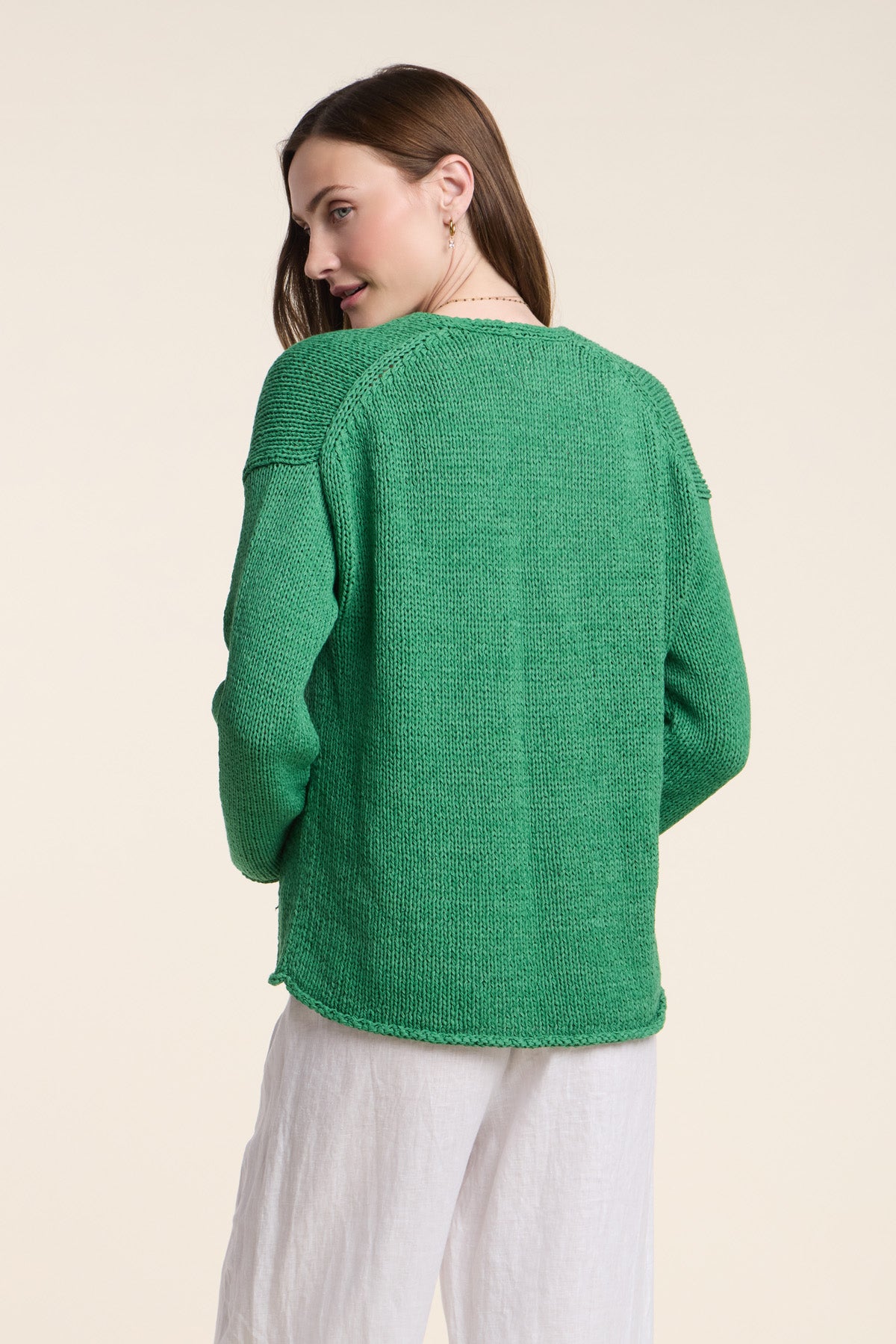 A woman faces away, wearing the QUINNLY CARDIGAN by Velvet by Graham & Spencer—an open-front green piece in cotton tape yarn—with white pants against a plain light background.