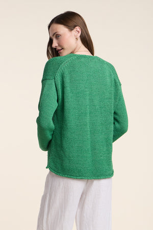 A woman faces away, wearing the QUINNLY CARDIGAN by Velvet by Graham & Spencer—an open-front green piece in cotton tape yarn—with white pants against a plain light background.