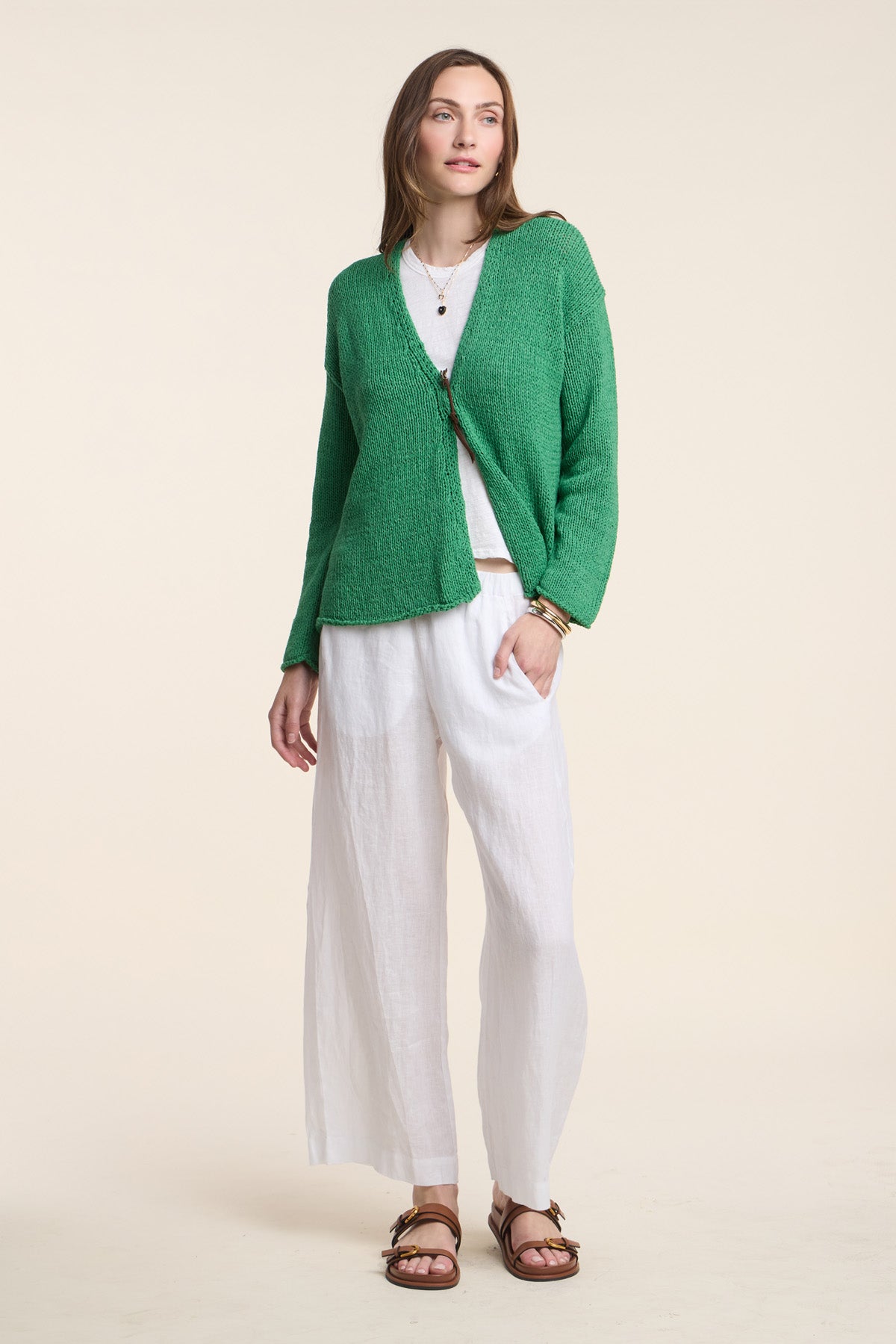 A woman stands against a plain light background wearing the LOLA LINEN PANT by Velvet by Graham & Spencer, styled with a green cardigan, white top, and brown sandals.