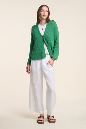 A woman stands against a plain light background wearing the LOLA LINEN PANT by Velvet by Graham & Spencer, styled with a green cardigan, white top, and brown sandals.
